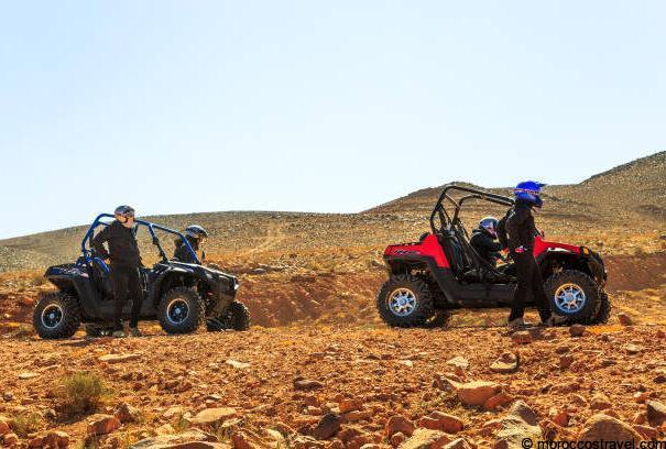 Moto Quad Bike Excursions & Tours In Desert - Merzouga Merzouga, Morocco - February 21, 2016: Four riders from two quad ATVs stand beside their vehicles on rocky terrain and survey the path ahead. Overhead is an overcast sky and in the distance a range of large hills can be seen